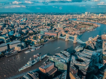 An aerial view of a bustling cityscape featuring a prominent river cutting through the landscape. Tower Bridge, with its iconic architecture, spans the river, surrounded by a mix of modern and historic buildings. The scene is under a blue sky punctuated by a few clouds, with sunlight casting warm tones over the city.