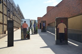 An outdoor art installation features life-sized vertical mirrors with images of people walking away, creating an optical illusion of figures in motion. The setting includes a concrete walkway lined with tall brick and stone walls, accentuated by clear blue skies.