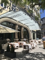 Outdoor dining area of a restaurant with elegant architecture. Tables are set with white tablecloths and placed under a glass canopy. A few patrons are seen walking by, enjoying the sunny day. The area is adorned with ornamental elements and small potted plants.