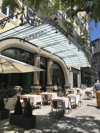 Outdoor dining area of a restaurant with elegant architecture. Tables are set with white tablecloths and placed under a glass canopy. A few patrons are seen walking by, enjoying the sunny day. The area is adorned with ornamental elements and small potted plants.
