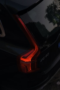 A dark vehicle with illuminated rear taillights, positioned diagonally with visible red and orange lighting detail. The car model is marked 'XC 90' and part of the surrounding environment, possibly trees, is reflected on the car's surface, adding to its sleek appearance.