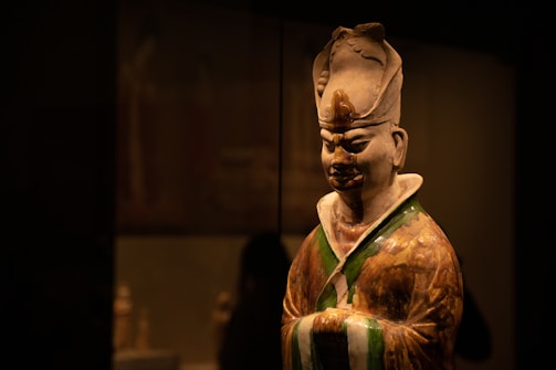 A detailed sculpture of an ancient figure wearing traditional attire with a headdress, in an art gallery setting. The lighting is focused on the sculpture, creating a dramatic contrast against the dark background.