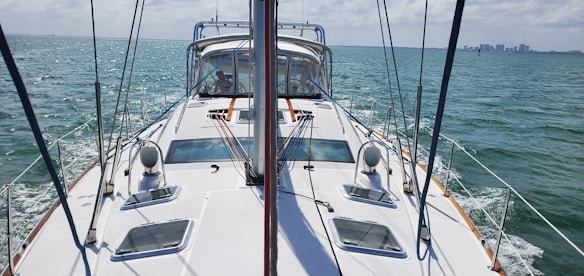 The image provides a view from the deck of a sailboat navigating through calm waters. The boat is equipped with various ropes and pulleys, and features a large cockpit with a transparent cover. In the distance, a skyline with several buildings is visible across the water.