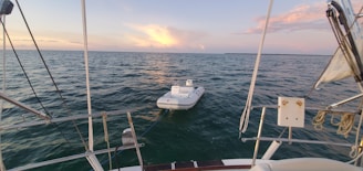 A small inflatable boat is tethered to the back of a larger sailboat, floating on a calm sea under a picturesque sky with scattered clouds and a setting sun casting a warm glow.