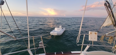 A small inflatable boat is tethered to the back of a larger sailboat, floating on a calm sea under a picturesque sky with scattered clouds and a setting sun casting a warm glow.
