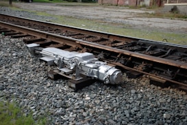 A mechanical apparatus installed next to railway tracks on a bed of gravel, possibly part of a railway turnout or switch system. The background shows greenery and a building with a paved area nearby.