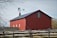 A large red barn with a metal roof is situated behind a wooden fence. A weather vane sits on top, with bare tree branches framing the scene. The barn has several small windows and a distinctively shaped roofline.