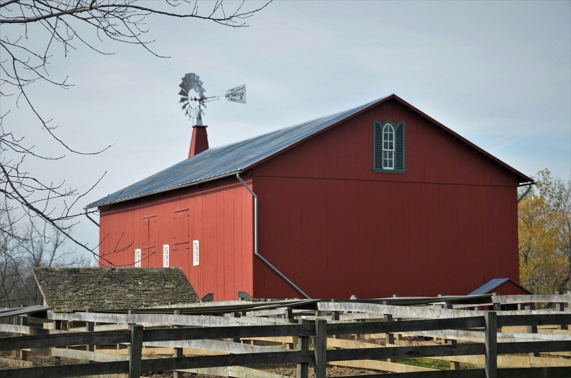 A large red barn with a metal roof is situated behind a wooden fence. A weather vane sits on top, with bare tree branches framing the scene. The barn has several small windows and a distinctively shaped roofline.