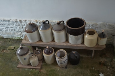 A collection of ceramic jugs and pots arranged on a wooden bench. The jugs are of varying sizes and feature a traditional design with handled lids. They are placed on a weathered wooden surface against a white stone wall and rough, mossy floor.