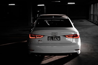 A white Audi car is parked indoors in a dimly lit space, with its rear lights illuminated and a license plate that reads 'ELIA24'. The Audi logo is prominently visible, and the setting gives an impression of a parking garage with dim artificial lighting.