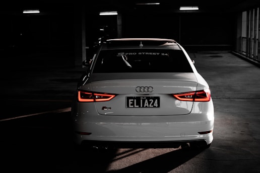 A white Audi car is parked indoors in a dimly lit space, with its rear lights illuminated and a license plate that reads 'ELIA24'. The Audi logo is prominently visible, and the setting gives an impression of a parking garage with dim artificial lighting.