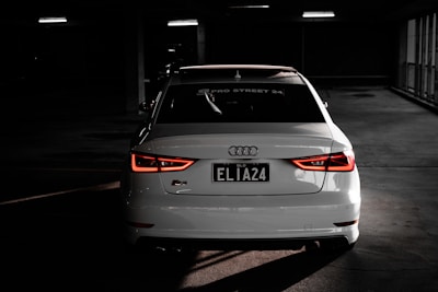 A white Audi car is parked indoors in a dimly lit space, with its rear lights illuminated and a license plate that reads 'ELIA24'. The Audi logo is prominently visible, and the setting gives an impression of a parking garage with dim artificial lighting.