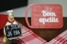A cheerful chef figurine holds a sign with the message 'A DIETA J&Aacute; ERA,' which translates to 'The diet is over.' In the background, a red cutting board features the phrase 'Bom apetite' with a chef's hat graphic. The scene includes a red and white checkered tablecloth.