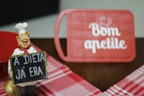 A cheerful chef figurine holds a sign with the message 'A DIETA J&Aacute; ERA,' which translates to 'The diet is over.' In the background, a red cutting board features the phrase 'Bom apetite' with a chef's hat graphic. The scene includes a red and white checkered tablecloth.