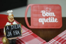 A cheerful chef figurine holds a sign with the message 'A DIETA J&Aacute; ERA,' which translates to 'The diet is over.' In the background, a red cutting board features the phrase 'Bom apetite' with a chef's hat graphic. The scene includes a red and white checkered tablecloth.