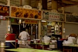 A food stall in a market features signs advertising Hainanese chicken rice, kaya toast, and other local dishes. The stall is equipped with cooking utensils, a counter, and storage shelves stocked with supplies. A person, likely a staff member, is wearing a white t-shirt and working at the counter. There are plastic containers and bowls on green-topped tables in the foreground.