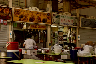 A food stall in a market features signs advertising Hainanese chicken rice, kaya toast, and other local dishes. The stall is equipped with cooking utensils, a counter, and storage shelves stocked with supplies. A person, likely a staff member, is wearing a white t-shirt and working at the counter. There are plastic containers and bowls on green-topped tables in the foreground.
