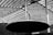 A monochrome architectural detail featuring a complex arrangement of metal beams and a large circular disc. The structure shows a pattern of perforated panels, creating a futuristic feel.