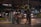 A nighttime urban scene with heavy traffic including cars and motorbikes. A person wearing a helmet stands near a bicycle and looks at something in their hands. Streetlights and headlights illuminate the area, while a nearby building with visible signage has decorative lighting.
