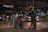 A nighttime urban scene with heavy traffic including cars and motorbikes. A person wearing a helmet stands near a bicycle and looks at something in their hands. Streetlights and headlights illuminate the area, while a nearby building with visible signage has decorative lighting.