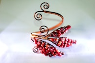 An intricately designed wire bracelet featuring swirling copper wires and clusters of red beads arranged in a floral pattern.