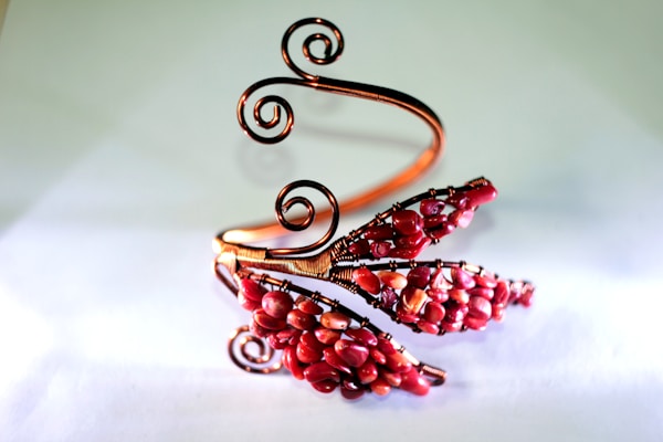 An intricately designed wire bracelet featuring swirling copper wires and clusters of red beads arranged in a floral pattern.