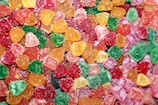 A close-up view of an assortment of colorful gummy candies shaped like small hearts. The candies are coated with a layer of sugar, giving them a slightly frosted appearance. The vibrant colors include red, green, orange, and yellow.