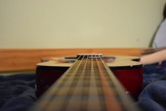 An acoustic guitar is positioned horizontally on a dark, soft surface with a focus on the strings stretching into the distance. The body of the guitar is visible with a reddish wood finish and is aligned against a light green wall.