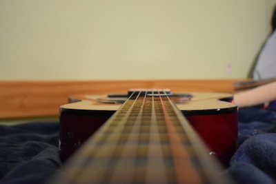 An acoustic guitar is positioned horizontally on a dark, soft surface with a focus on the strings stretching into the distance. The body of the guitar is visible with a reddish wood finish and is aligned against a light green wall.