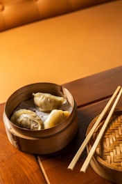 Three dumplings are placed inside a bamboo steamer on a wooden table. Two chopsticks rest beside the steamer on the table. The background features a light brown cushion, enhancing the warm ambiance.