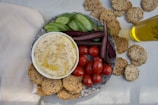 A stone plate holds a bowl of creamy hummus garnished with olive oil, surrounded by sliced cucumbers, purple carrots, and cherry tomatoes. Seeded crackers are arranged around the plate on a light-colored cloth. A bottle of olive oil is placed nearby, along with more scattered crackers.