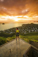 A person stands on a concrete platform overlooking a scenic view of a coastline at sunset. The sky is filled with dramatic orange clouds, and the sun is setting over the ocean. Below, a lush green landscape with hills and a small town can be seen. On the platform, there is graffiti that includes the word 'HAWAII.'