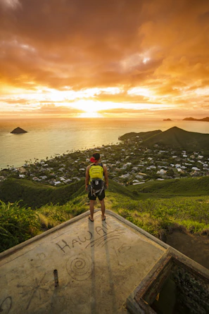 A person stands on a concrete platform overlooking a scenic view of a coastline at sunset. The sky is filled with dramatic orange clouds, and the sun is setting over the ocean. Below, a lush green landscape with hills and a small town can be seen. On the platform, there is graffiti that includes the word 'HAWAII.'