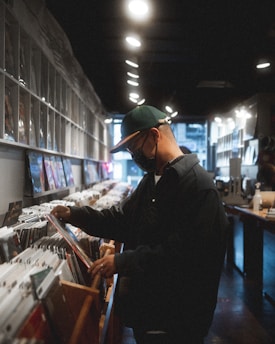 A person wearing a cap and mask is browsing through vinyl records in a dimly lit record store. Shelves filled with record covers stretch along the walls, and overhead lights cast a soft glow.