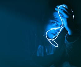 A person holds a glowing blue neon light close to their face, creating a dramatic and moody illumination. The surrounding environment is dark, emphasizing the striking contrast between light and shadow.
