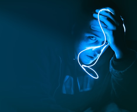 A person holds a glowing blue neon light close to their face, creating a dramatic and moody illumination. The surrounding environment is dark, emphasizing the striking contrast between light and shadow.