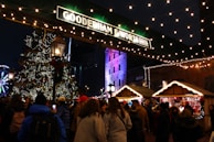 A festive scene in a bustling market at night, featuring a large Christmas tree adorned with lights and ornaments. Many people are gathered, dressed in winter clothing, surrounded by wooden stalls selling holiday items. Strings of lights are hung overhead, adding warmth and charm. A Ferris wheel is visible in the background, and a historic building is illuminated with colorful lights.