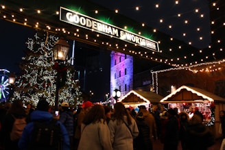 A festive scene in a bustling market at night, featuring a large Christmas tree adorned with lights and ornaments. Many people are gathered, dressed in winter clothing, surrounded by wooden stalls selling holiday items. Strings of lights are hung overhead, adding warmth and charm. A Ferris wheel is visible in the background, and a historic building is illuminated with colorful lights.