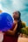 A young woman with long braided hair holds a blue balloon with the words 'Happy Birthday' printed on it. She is wearing a burgundy dress and standing outdoors with a clear sky and neatly trimmed trees in the background.