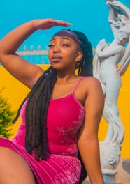 A woman with long black braided hair is posing outdoors against a vibrant yellow background and clear blue sky, with a white statue to her right. She is wearing a bright pink sleeveless dress and appears to be shielding her eyes from the sun with her hand on her forehead.