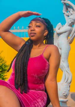A woman with long black braided hair is posing outdoors against a vibrant yellow background and clear blue sky, with a white statue to her right. She is wearing a bright pink sleeveless dress and appears to be shielding her eyes from the sun with her hand on her forehead.