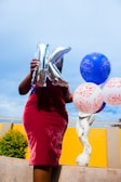 A person holding a silver balloon shaped like the letter 'K' and several colorful balloons, some with decorative patterns. The background includes a yellow wall and a sculpture of a cherub. The person is wearing a maroon velvet dress.
