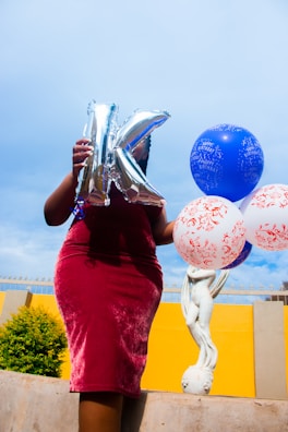 A person holding a silver balloon shaped like the letter 'K' and several colorful balloons, some with decorative patterns. The background includes a yellow wall and a sculpture of a cherub. The person is wearing a maroon velvet dress.