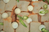Square pieces of a light-colored, slightly speckled baked good are topped with small spherical white objects resembling eggs. Scattered among them are slices of fresh green chili peppers, adding vibrant green accents.
