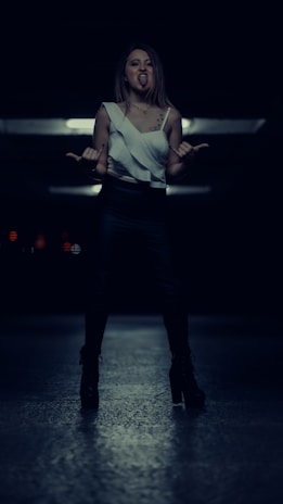 A person stands confidently in a dimly lit environment, wearing dark pants and a sleeveless top. The individual is making a playful gesture with their hands, sticking out their tongue, and seeming to express attitude.