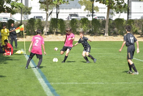 A group of young soccer players are actively engaged in a match. One player in a black uniform is challenging another player in a pink uniform for the ball in the central area of the field. There are additional players and a referee standing on the sideline near a corner flag. In the background, parked vehicles, a building, and trees are visible.