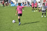 A child wearing a pink jersey with the number 10 prepares to kick a soccer ball on a grassy field. Other children in pink and black-and-white striped jerseys are in the background, suggesting a youth soccer match. Spectators can be seen in the distance.