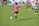 A child wearing a pink jersey with the number 10 prepares to kick a soccer ball on a grassy field. Other children in pink and black-and-white striped jerseys are in the background, suggesting a youth soccer match. Spectators can be seen in the distance.