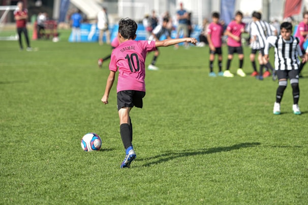 A child wearing a pink jersey with the number 10 prepares to kick a soccer ball on a grassy field. Other children in pink and black-and-white striped jerseys are in the background, suggesting a youth soccer match. Spectators can be seen in the distance.