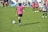 A child wearing a pink jersey with the number 10 prepares to kick a soccer ball on a grassy field. Other children in pink and black-and-white striped jerseys are in the background, suggesting a youth soccer match. Spectators can be seen in the distance.